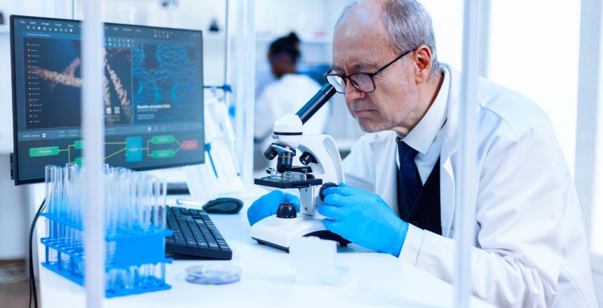 Senior scientist in chemistry laboratory uses microscope working on new medicine. Chemist researcher in sterile lab doing experiments for medical industry using modern technology.